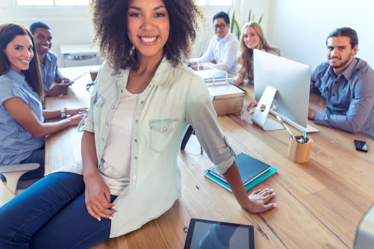 brown woman sitting on desk in conference room with her colleagues sitting behind her